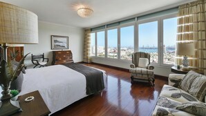 A hotel room with a large bed, a chair, a sofa, and a view of the city through large windows.
