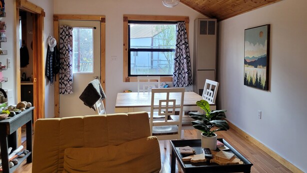 Un salon chaleureux avec un plafond en bois, une grande fenêtre avec des rideaux, une table à manger avec des chaises, un canapé et une table basse agrémentée d’objets décoratifs.
