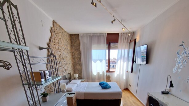 A compact bedroom with a bed, bedside table, and a desk with a chair. There is a window with curtains, a TV mounted on the wall, and a small plant on the floor. - Rooms Aribau 133 (Barcelona)
