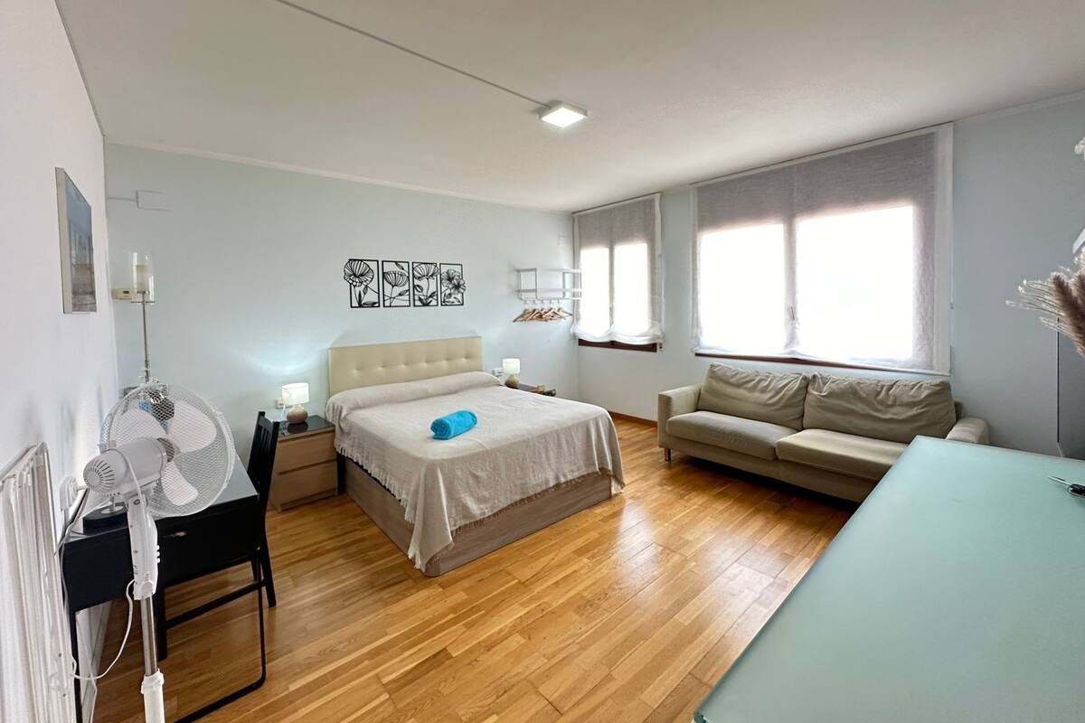 Ein Schlafzimmer mit Bett, Nachttisch, Lampe, Ventilator, Sofa und Fenster.
