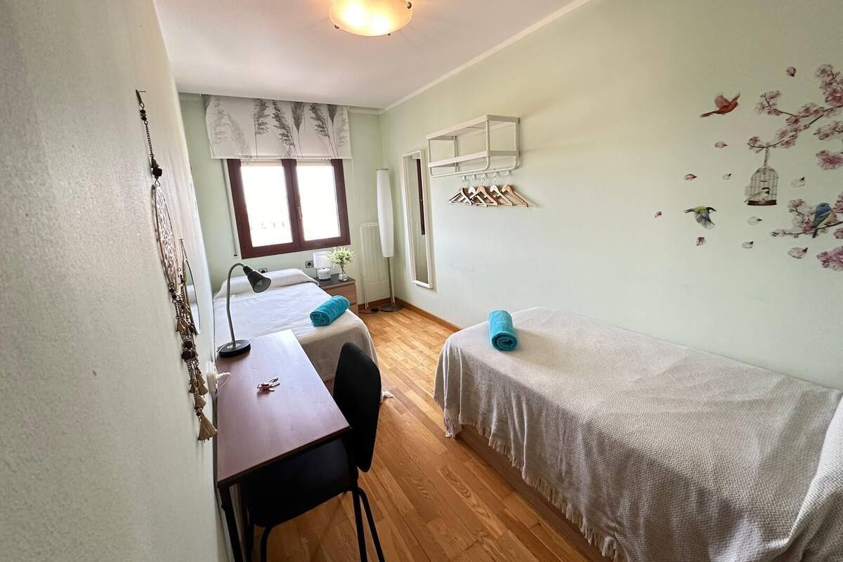 Ein kleiner, hell erleuchteter Raum mit einem Bett, einem Schreibtisch und einem Stuhl. Ein Fenster mit Vorhängen, eine Heizung und Wanddekorationen mit Vögeln und Blumen sind zu sehen.