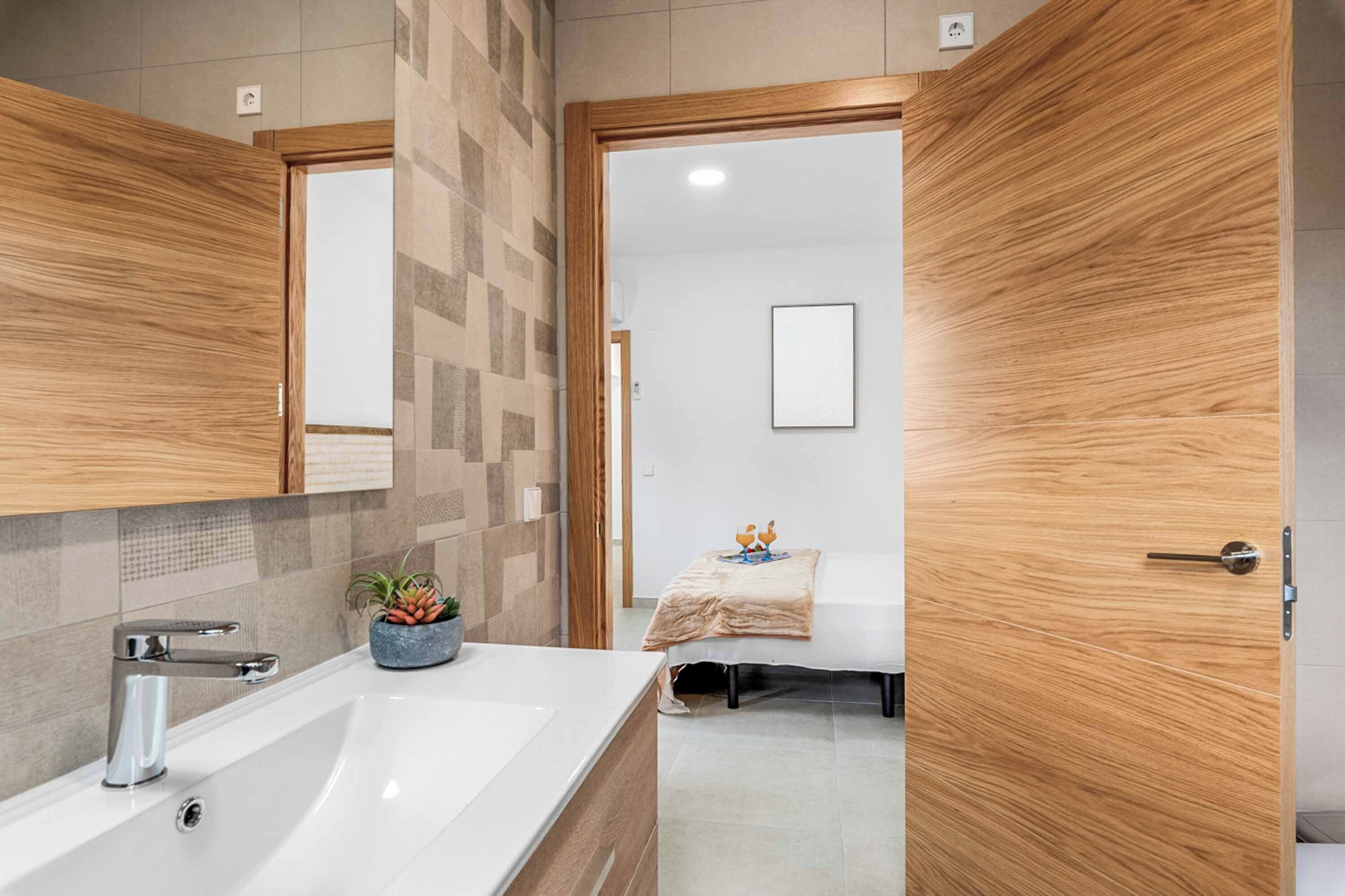 A modern bathroom with a large sink, wooden door, and a bed in the adjacent room.