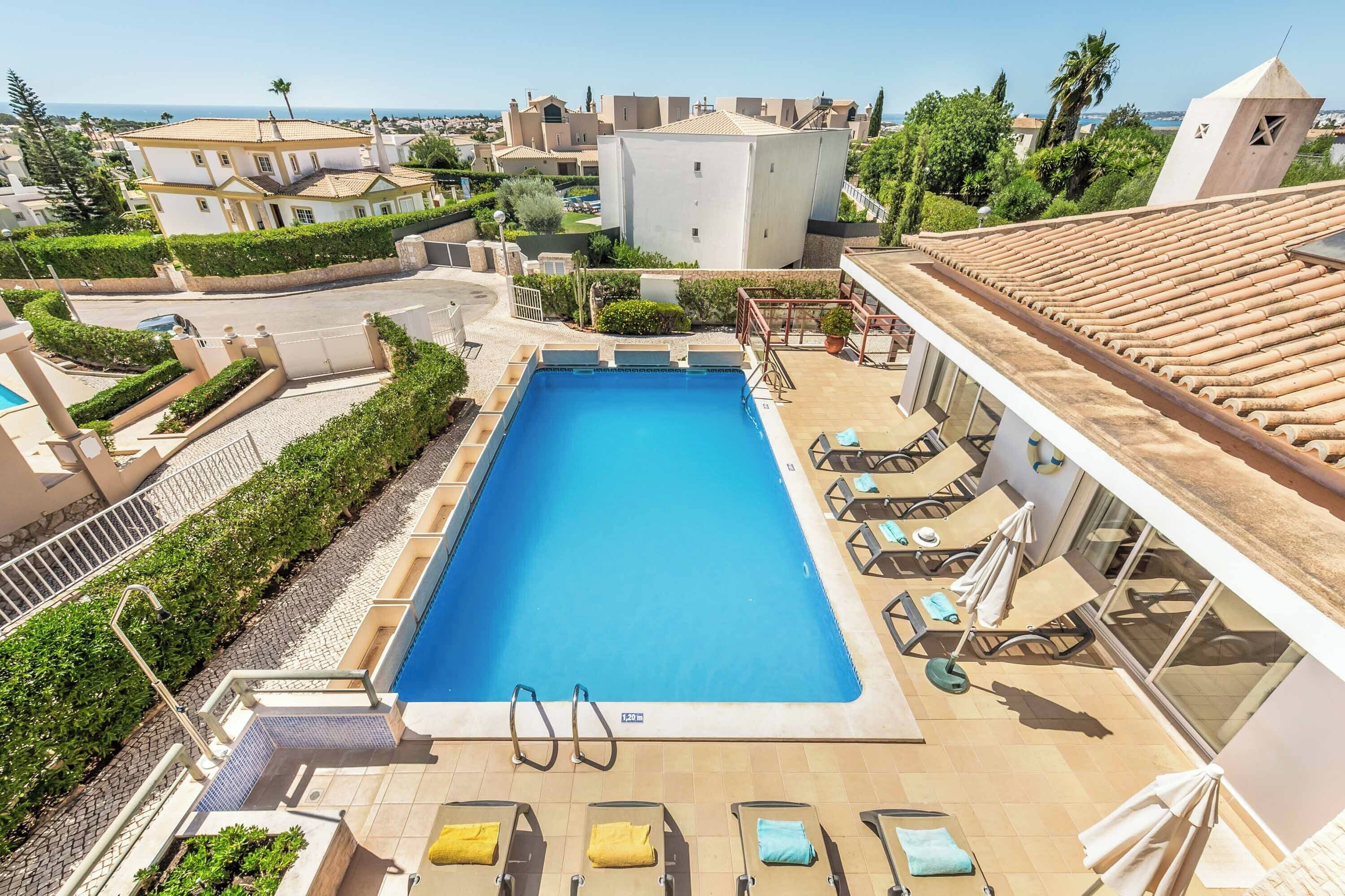 A swimming pool with lounge chairs and umbrellas, surrounded by greenery and buildings.