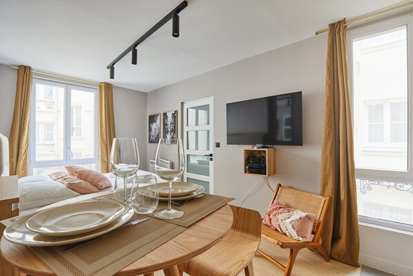 A hotel room with a dining table set for two, a television mounted on the wall, and a window with curtains.