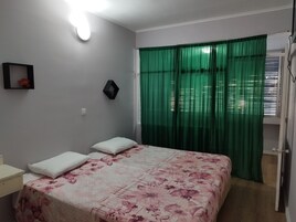 A bedroom with a bed, a nightstand, a wall-mounted shelf, and a window with green curtains.