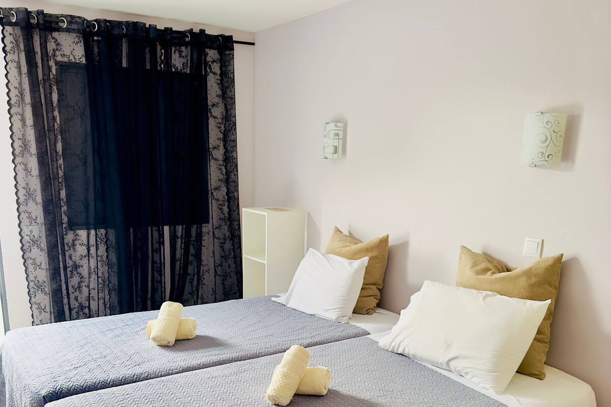 A double bed with grey bedding and two pillows, a bedside table, and a lamp.