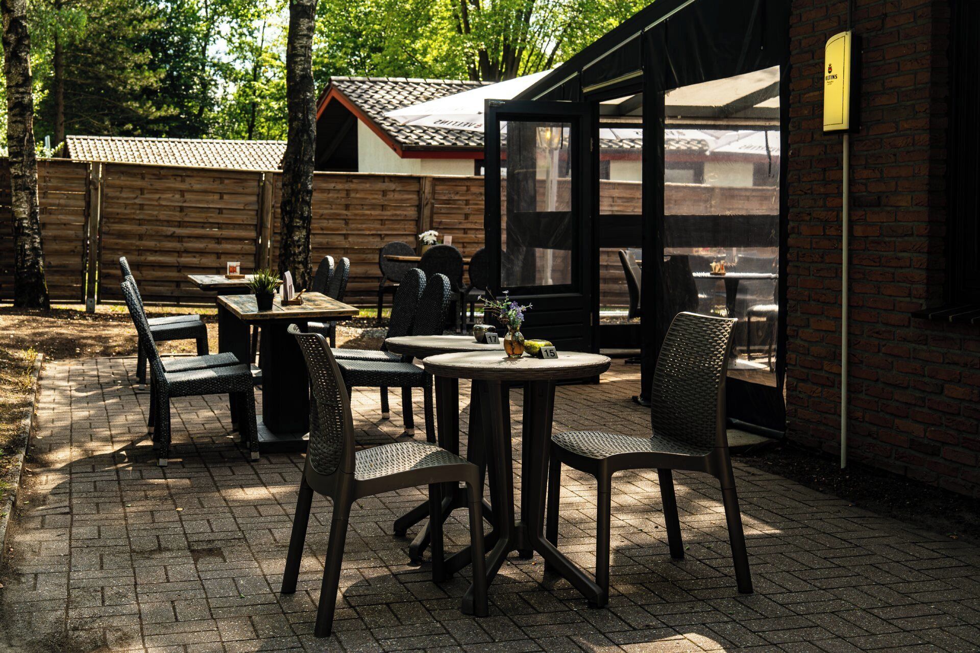 Een terras buiten met tafels en stoelen, een houten omheining en een bakstenen gebouw op de achtergrond.