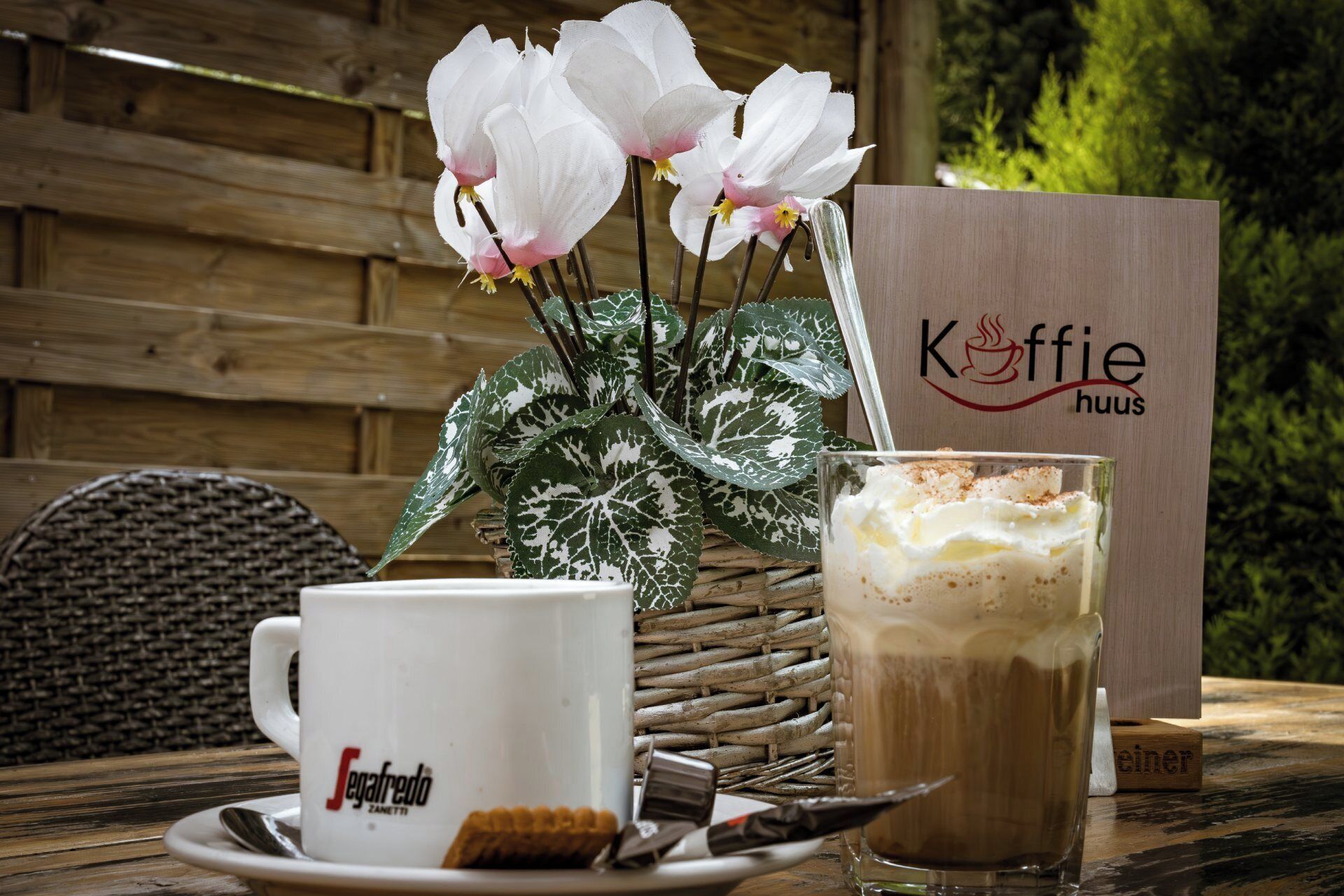 Una mesa con una taza blanca, una taza de café, un pastel y una planta decorativa.