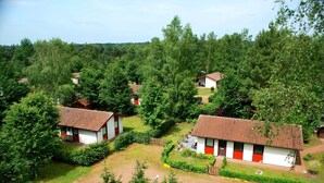 A cluster of white buildings with red accents, surrounded by dense greenery and a clear sky. - Type 6p - Two Bedroom Chalet, Sleeps 6 (Uelsen)