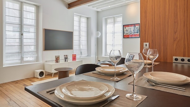 A dining area with a wooden table set for four, a television mounted on the wall, and large windows with blinds. - Amazing 2 Bedrooms Apartement Paris Center (Paris)