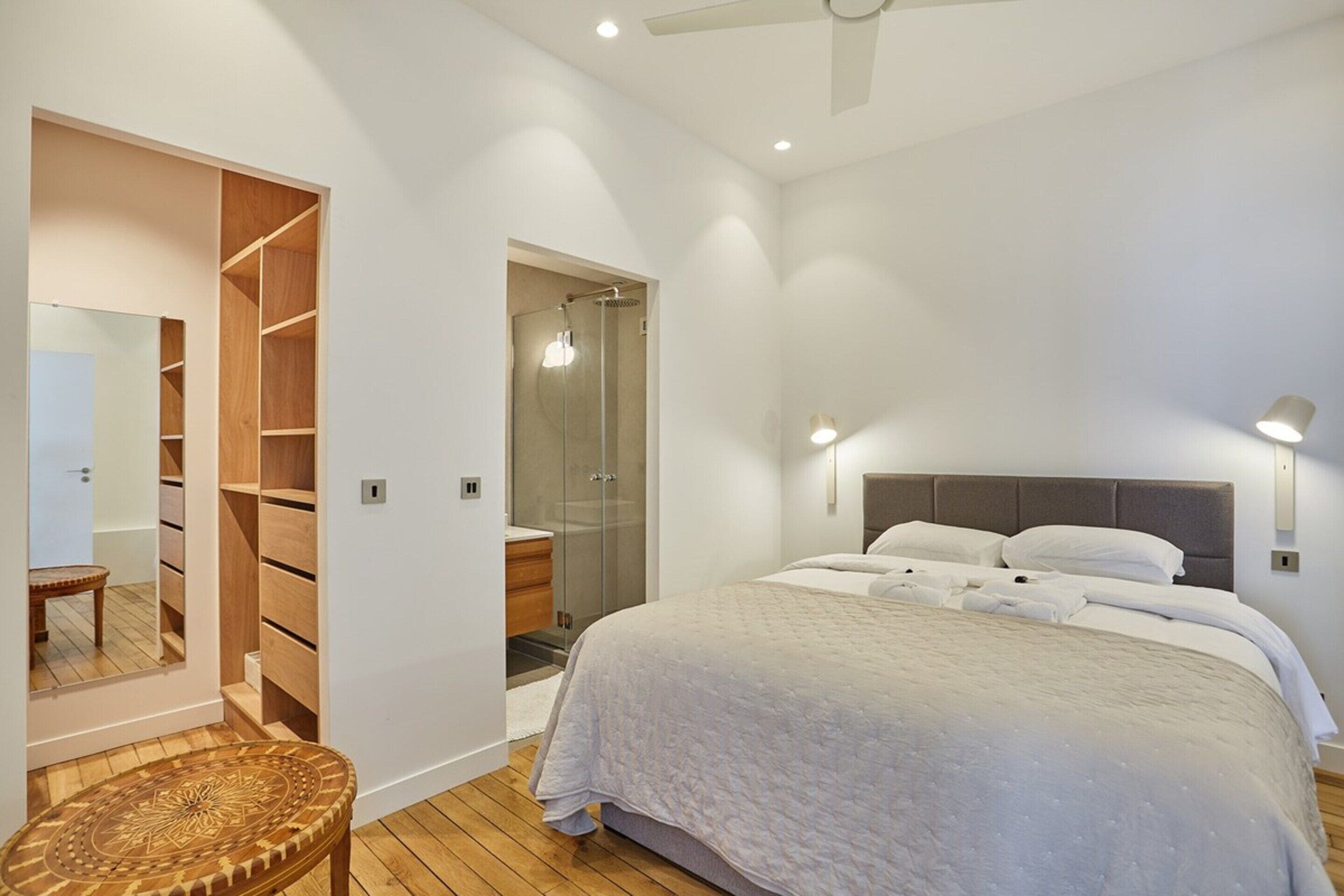 Un dormitorio con una cama grande, vestidor, ducha de vidrio y ventilador de techo.