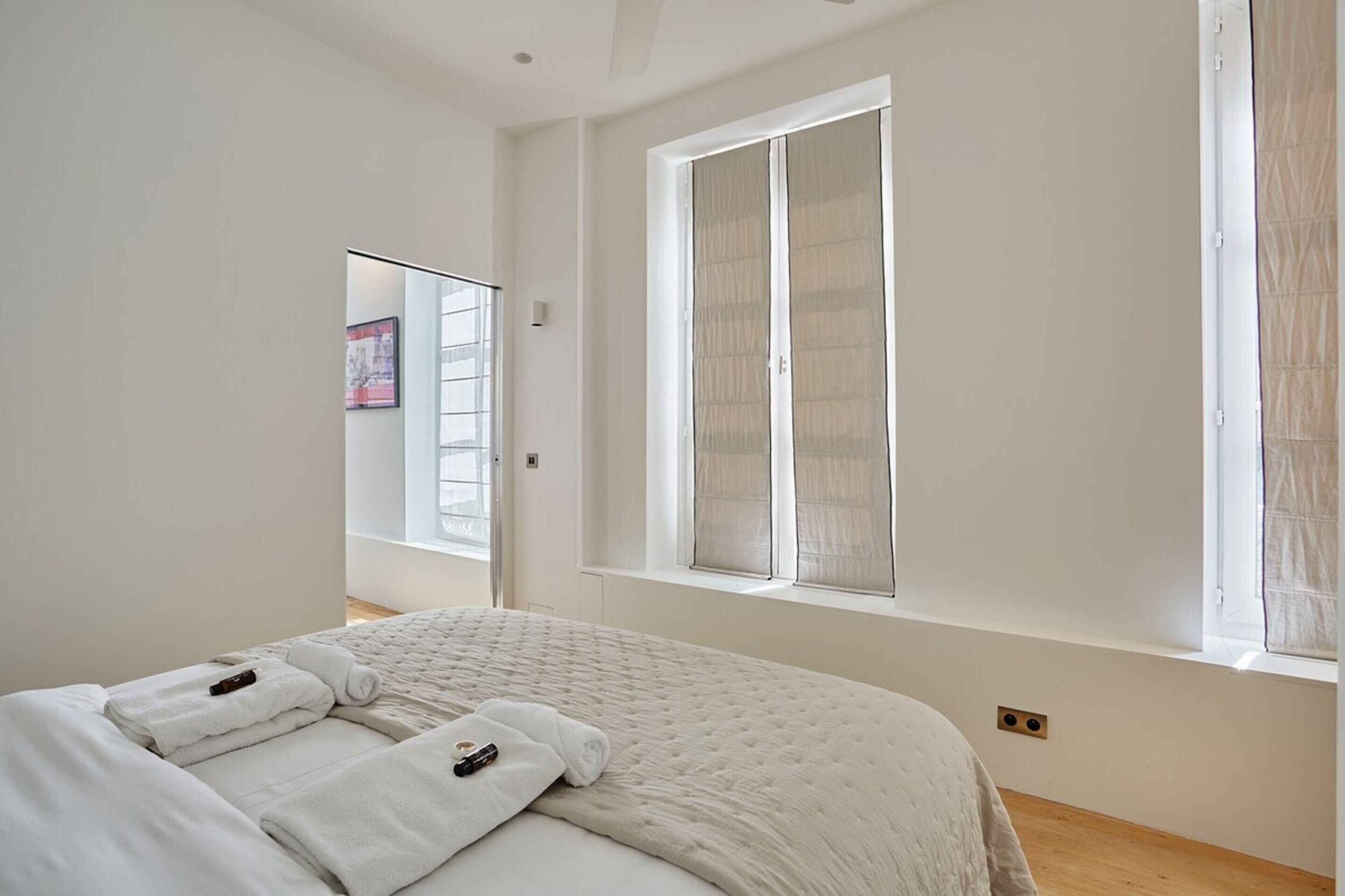A bedroom with a large bed, a window with blinds, and a door leading to another room.