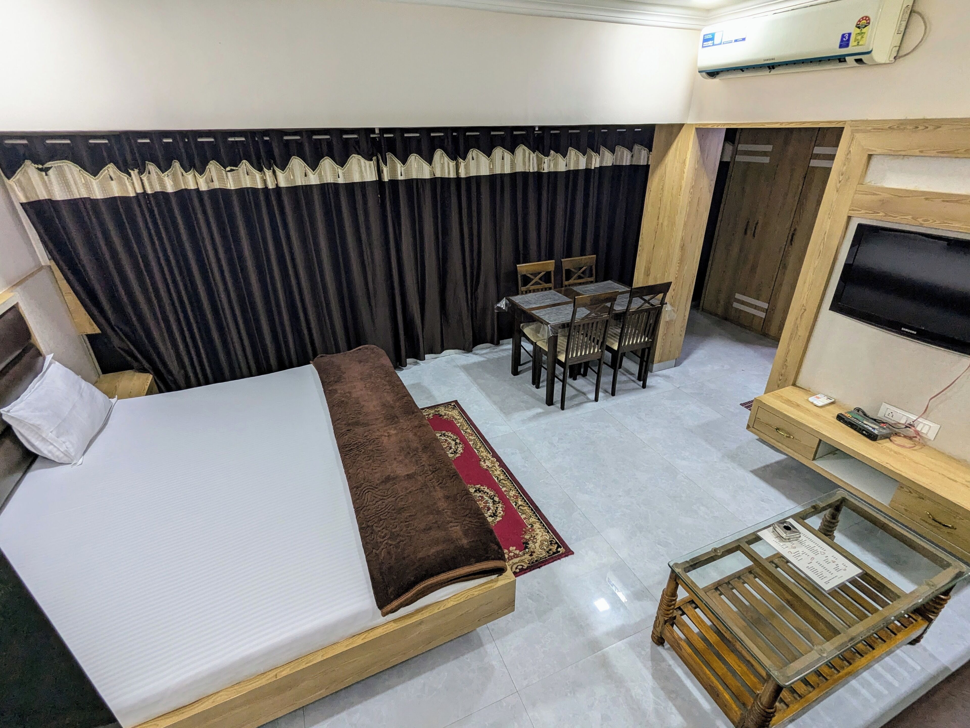 A hotel room with a bed, a desk with a TV, a chair, a small table, and a window with curtains.