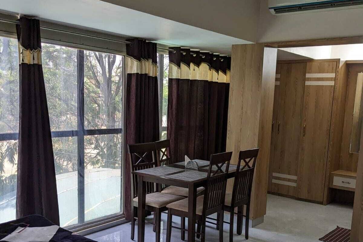A room with a dining table and chairs, a bed with a floral bedspread, and a large window with curtains.