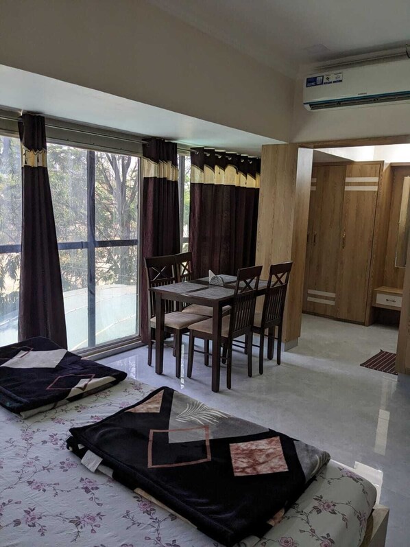 Um quarto com uma mesa de jantar e cadeiras, uma cama com colcha florida, e uma janela grande com cortinas.