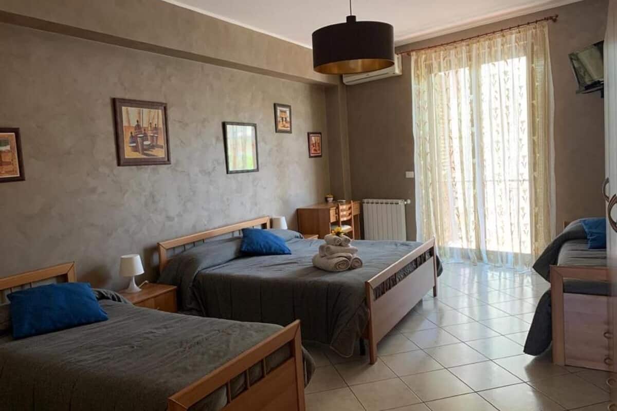 A hotel room with two single beds, a wooden headboard, a bedside table with a lamp, a radiator, and a window with sheer curtains.