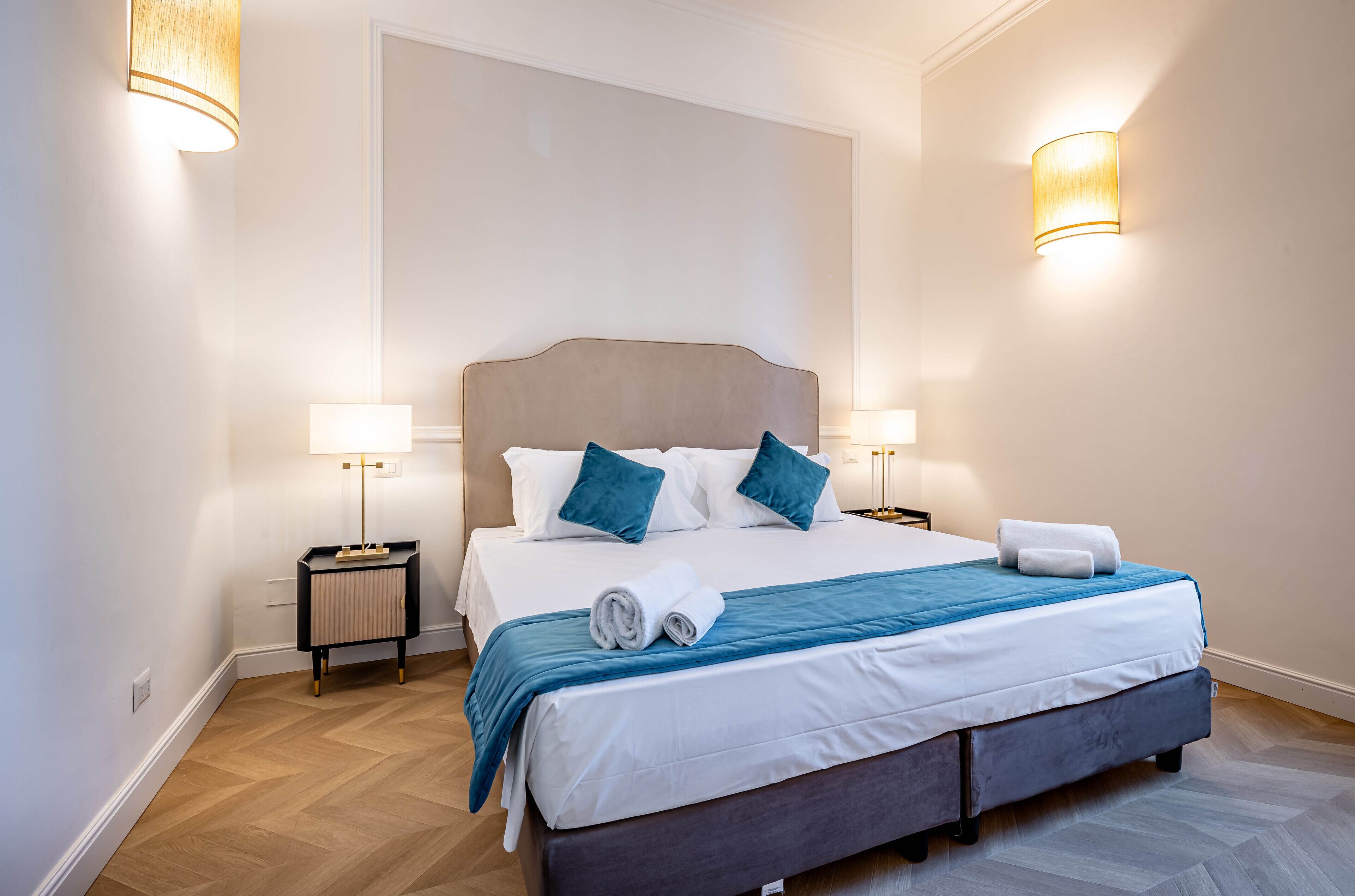 A neatly made bed with a blue blanket and pillows, a bedside table with a lamp, and a wall-mounted light fixture.