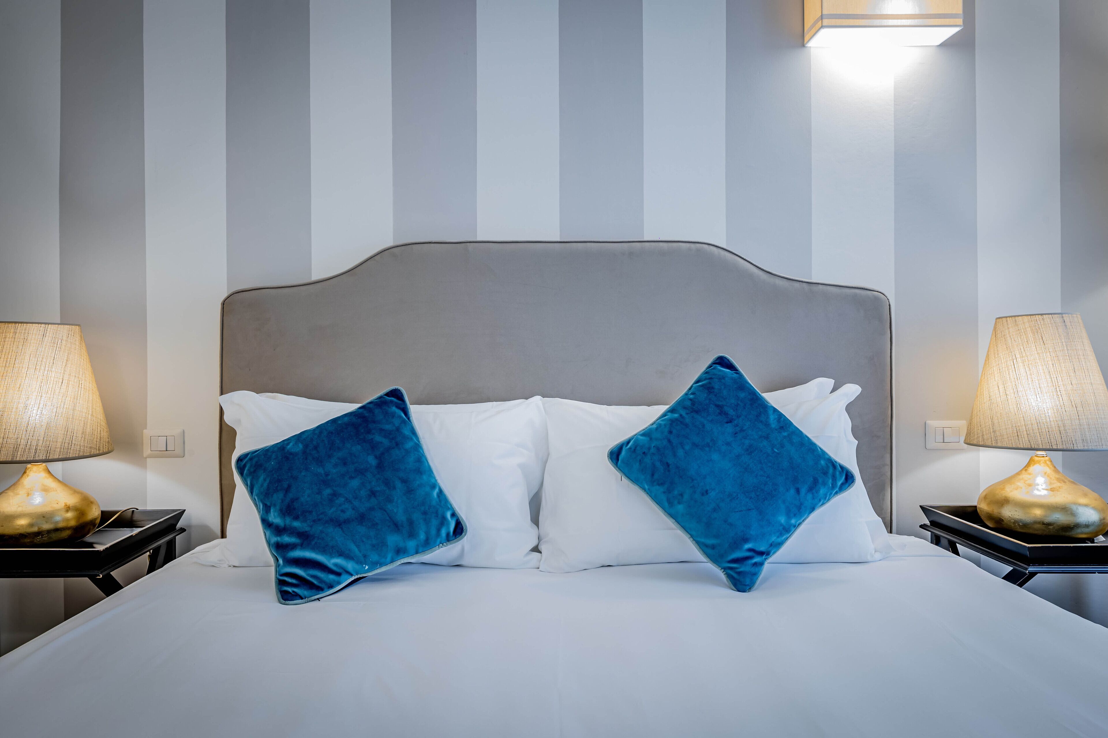 A neatly made bed with white linens and two blue pillows, flanked by two bedside lamps with beige shades.