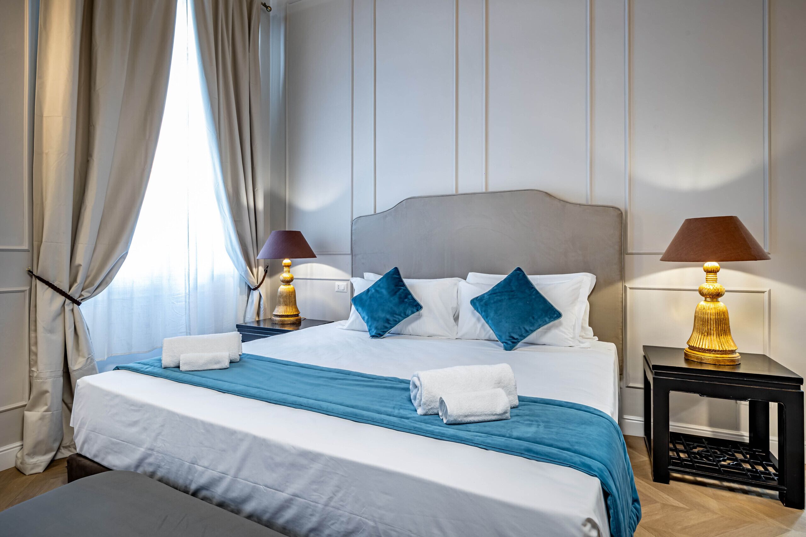 A neatly made bed with white and blue linens, two bedside lamps, and a window with curtains.