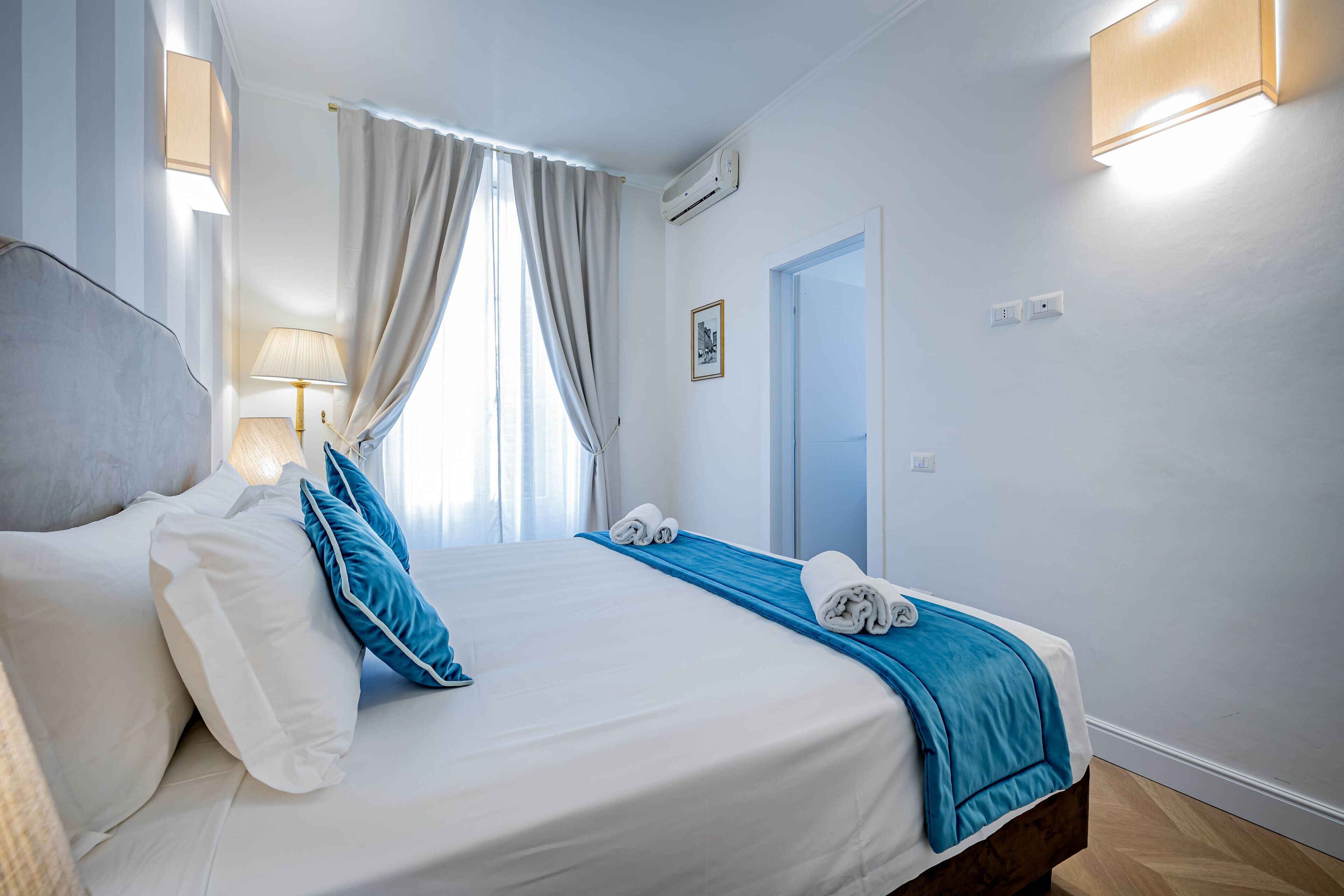 A neatly made bed with white and blue bedding, a bedside lamp, and a window with curtains.