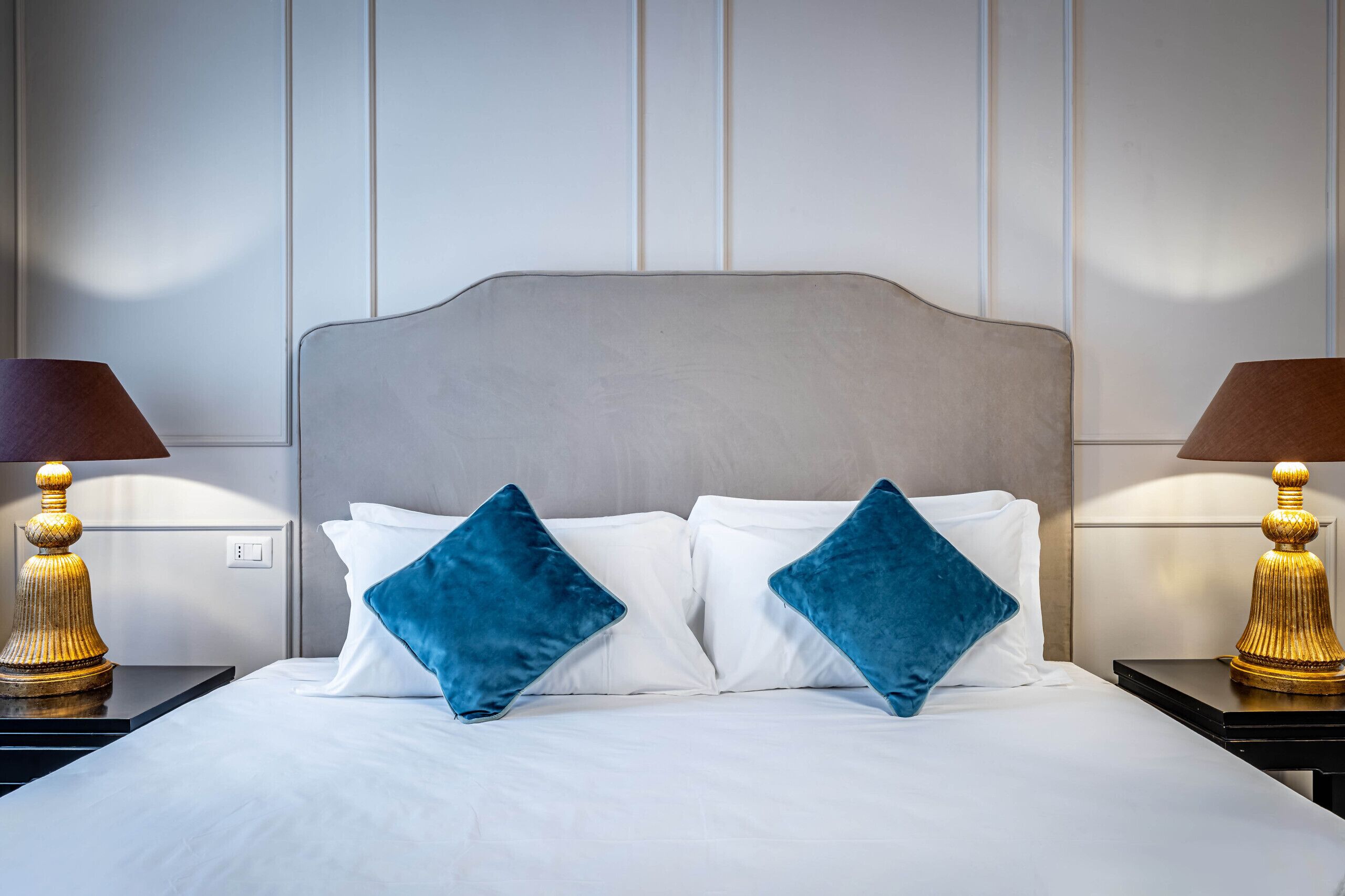 A neatly made bed with white linens and two blue pillows, flanked by two nightstands with lamps.