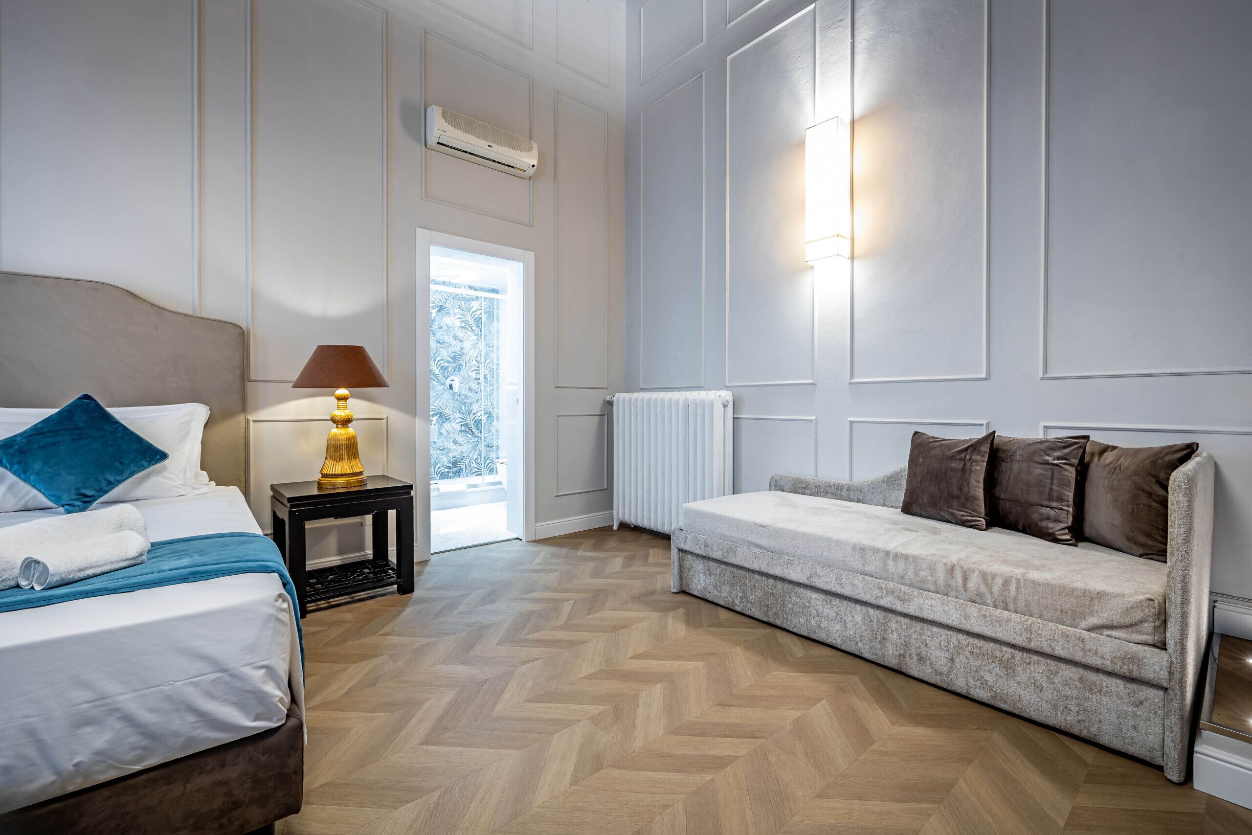 A hotel room with a bed, a sofa, a lamp, a radiator, and a window.