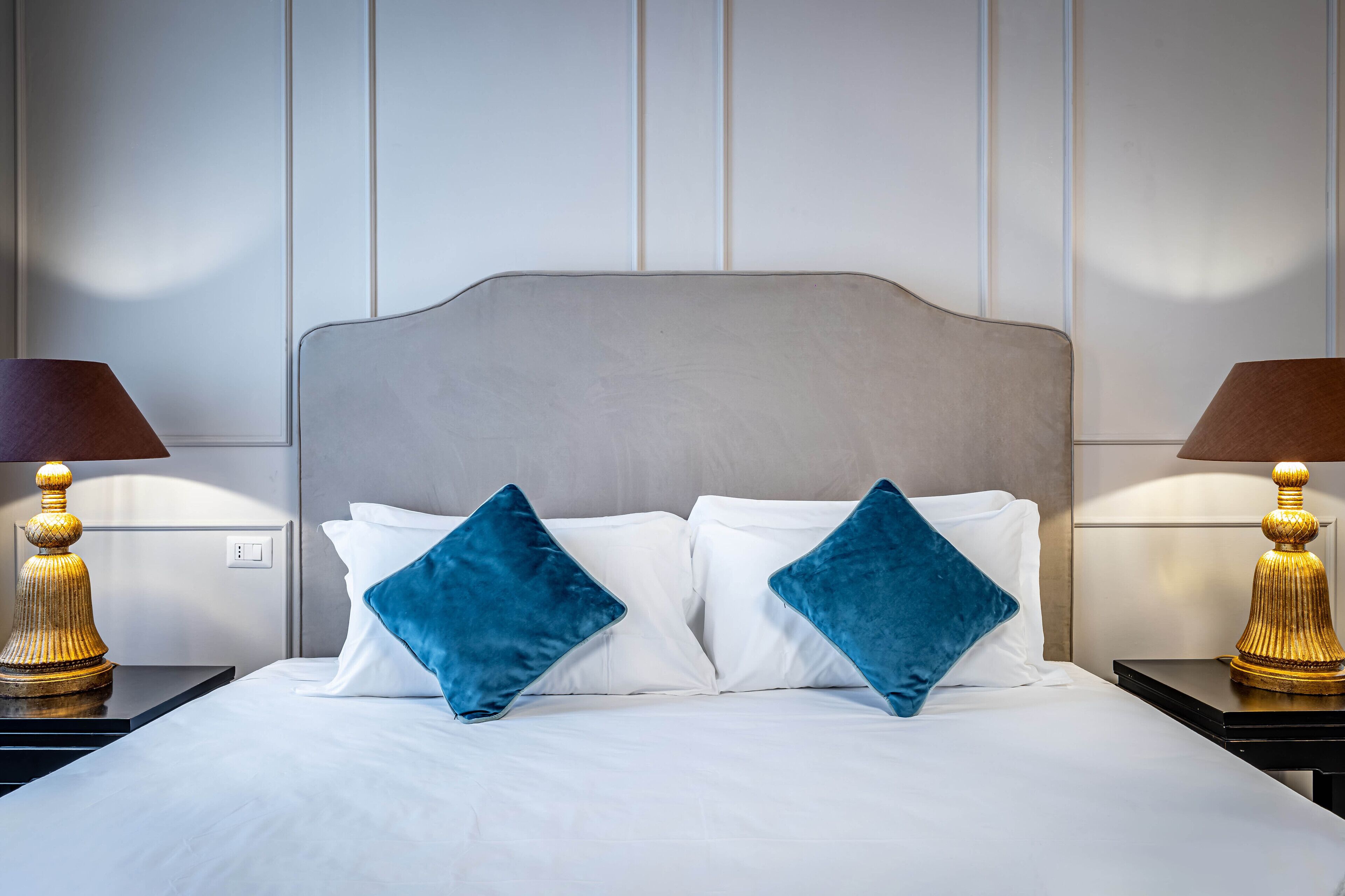 A neatly made bed with white linens and two blue pillows, flanked by two nightstands with lamps.