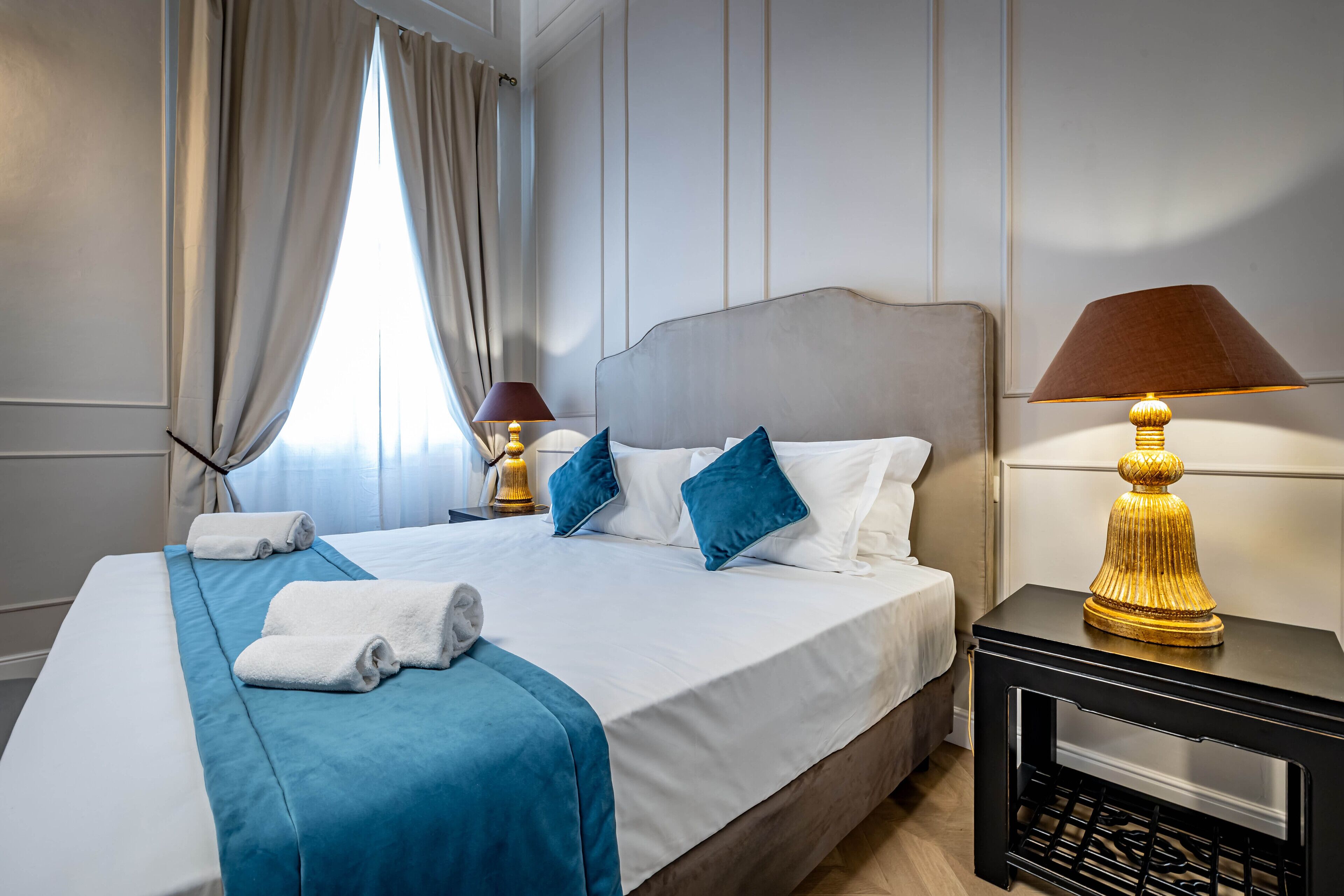 A neatly made bed with blue and white bedding, a gold lamp on a black nightstand, and a window with curtains.