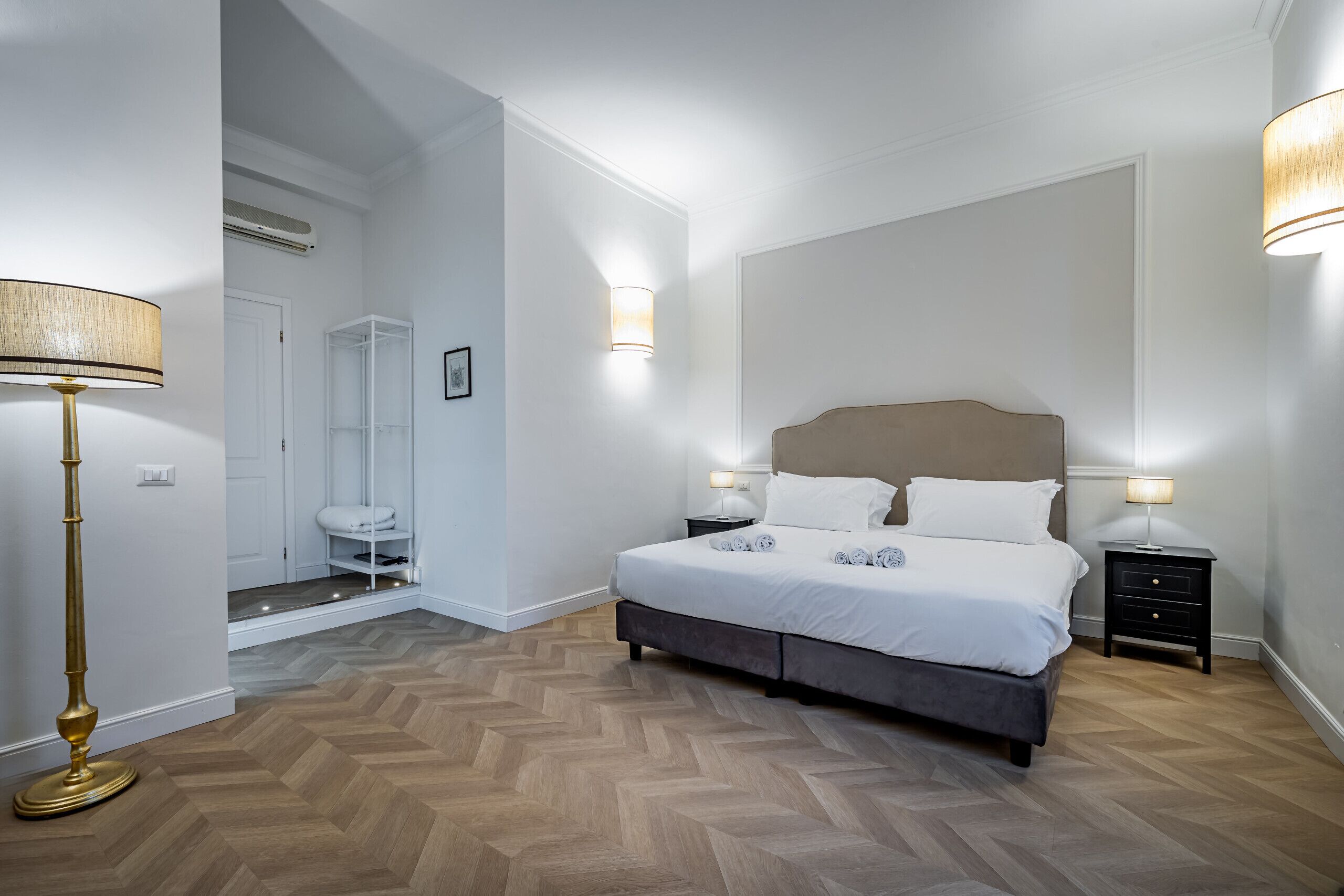 A bedroom with a bed, bedside tables, a floor lamp, a wardrobe, and a bathroom with a toilet and sink.