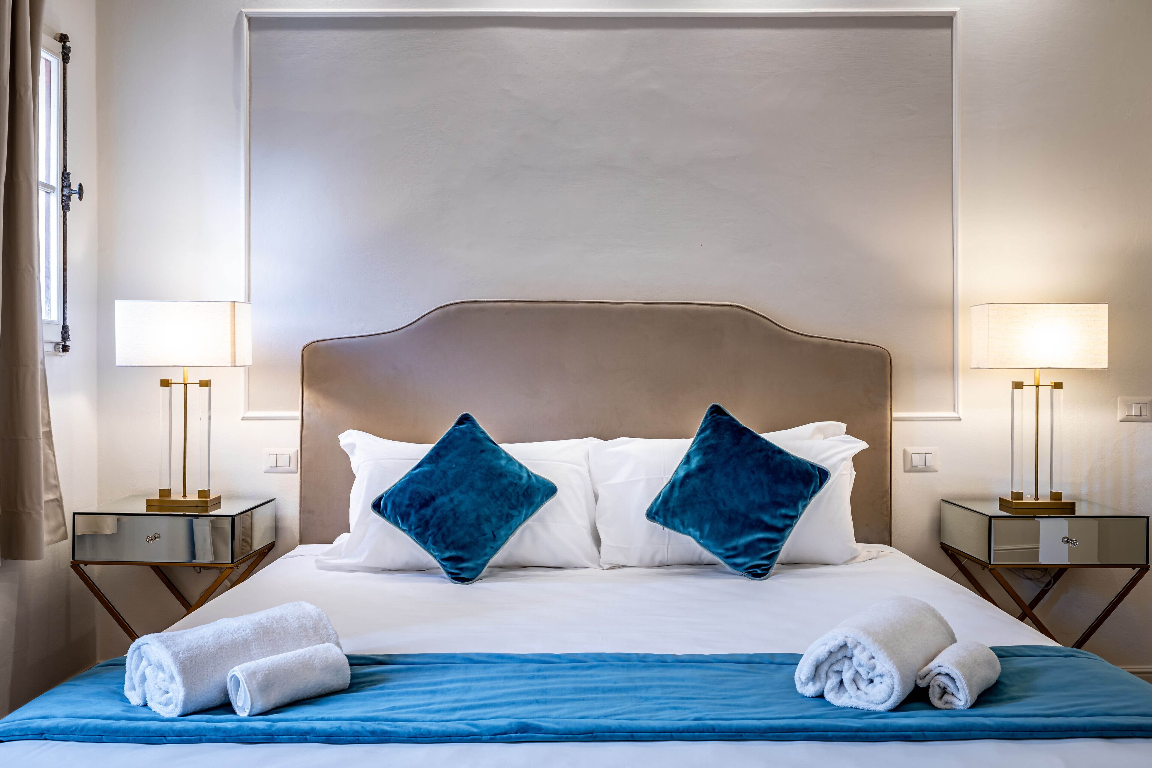 Un lit bien fait, avec du linge de lit blanc et des oreillers bleus, flanqué de deux tables de chevet sur lesquelles sont posées des lampes.