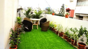 Un petit patio clos, agrémenté d’un gazon synthétique, d’une table ronde, de deux chaises en osier et de divers pots de plantes.