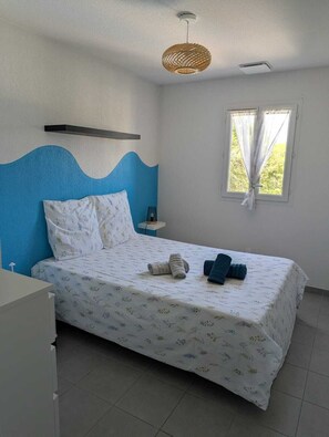 Ein Schlafzimmer mit Bett, Nachttisch, einem Fenster mit Vorhängen, einer Pendelleuchte und einer blauen Akzentwand.
