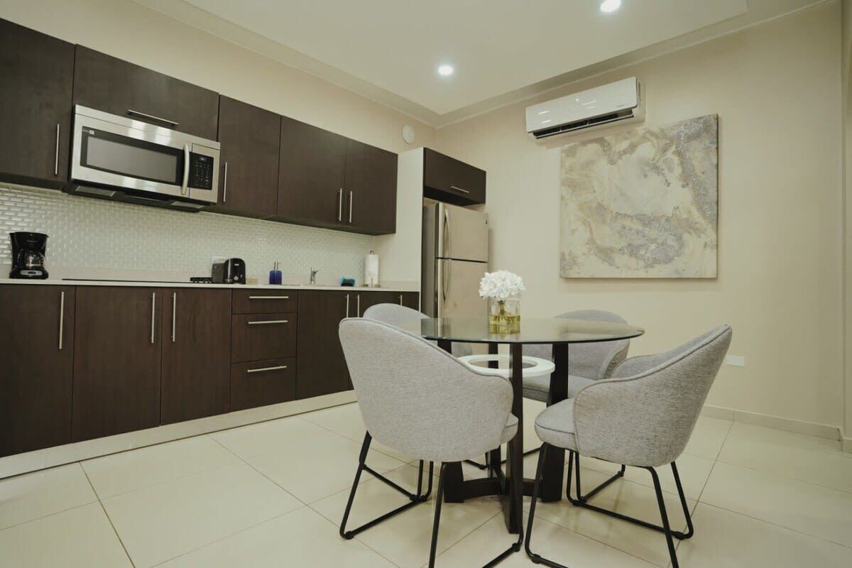 A modern kitchen with dark cabinets, a stainless steel microwave, a refrigerator, and a dining table with two chairs.