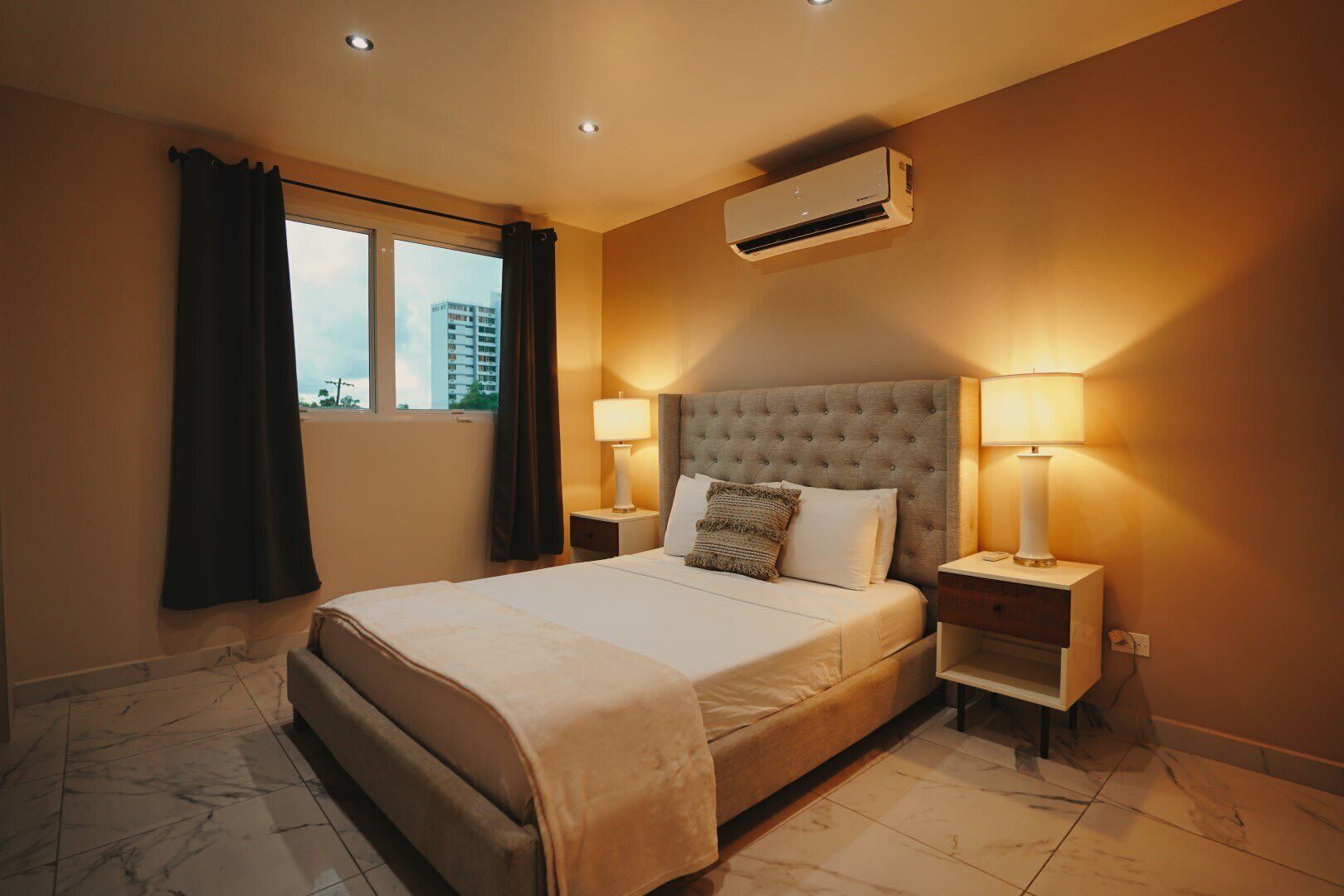 A hotel room with a large bed, two bedside lamps, an air conditioning unit, and a window with curtains.