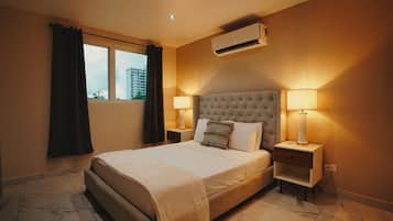 A hotel room with a large bed, two bedside lamps, an air conditioning unit, and a window with curtains.