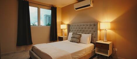 A hotel room with a large bed, two bedside lamps, an air conditioning unit, and a window with curtains.