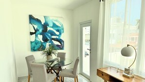 Un comedor moderno con una mesa de vidrio rodeada por cuatro sillas, un cuadro abstracto de gran tamaño en la pared, una planta en maceta y un escritorio de madera con una lámpara.