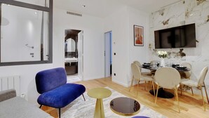 A modern living room with a blue sofa, a small round table, and a dining area with a TV mounted on the wall. - Authentic apt - 2br/6p - Rue des Martyrs - Opera (Paris)