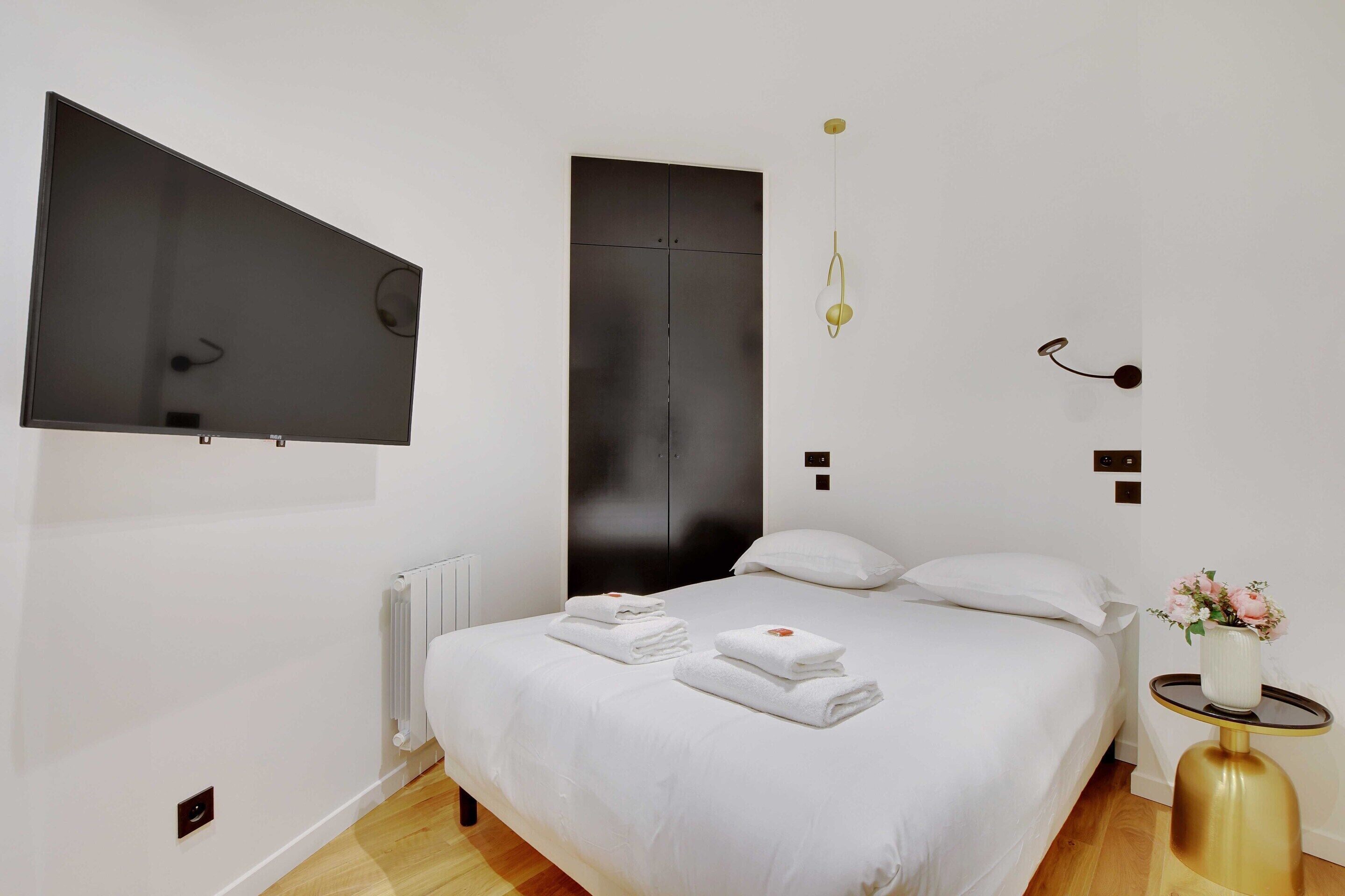 A modern hotel room with a large bed, a flat-screen TV mounted on the wall, a bedside table with a vase of flowers, and a black wardrobe.