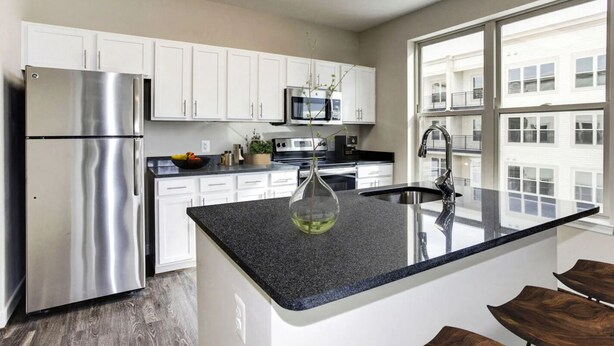 A modern kitchen with white cabinets, stainless steel appliances, a granite countertop, and a large window providing a view of the building's exterior. - National at Woodbridge (Woodbridge)