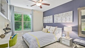 A bedroom with a bed, a desk, a chair, a nightstand, a lamp, and artwork on the wall. - National at Woodbridge (Woodbridge)