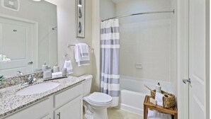 A bathroom with a granite countertop sink, a toilet, a shower with a striped curtain, and a wooden side table with toiletries. - National at Woodbridge (Woodbridge)