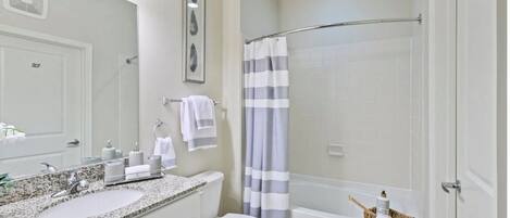 A bathroom with a granite countertop sink, a toilet, a shower with a striped curtain, and a wooden side table with toiletries.