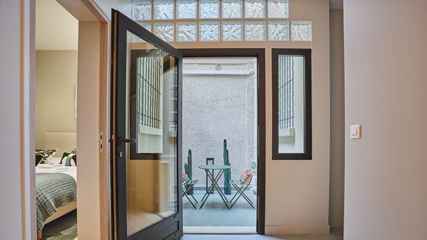 A hallway leading to a balcony with a table and chairs, a bed with a green bedspread, and a door with a frosted glass panel.