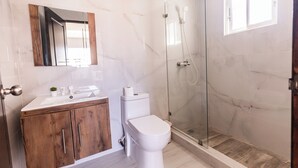 A bathroom with a white toilet, a wooden vanity, a glass shower enclosure, and a marble-tiled floor. - Hostal las Rosas de Punta Cana (Punta Cana)