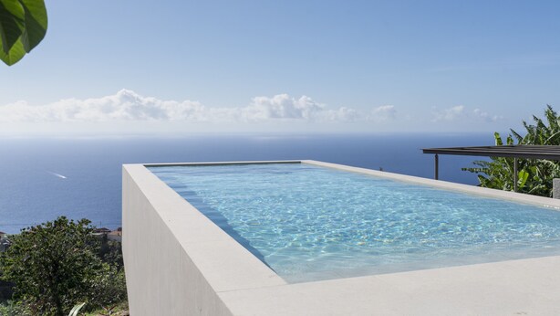 Uma piscina infinita retangular com água azul cristalina, rodeada por uma borda de concreto, com vista para a costa, o oceano e edifícios ao longe.