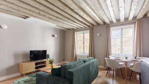 A modern living room with a sofa, TV, wooden flooring, and a dining area with chairs and a table. - Magnificent new Flat Le Marais/beaubourg (Paris)