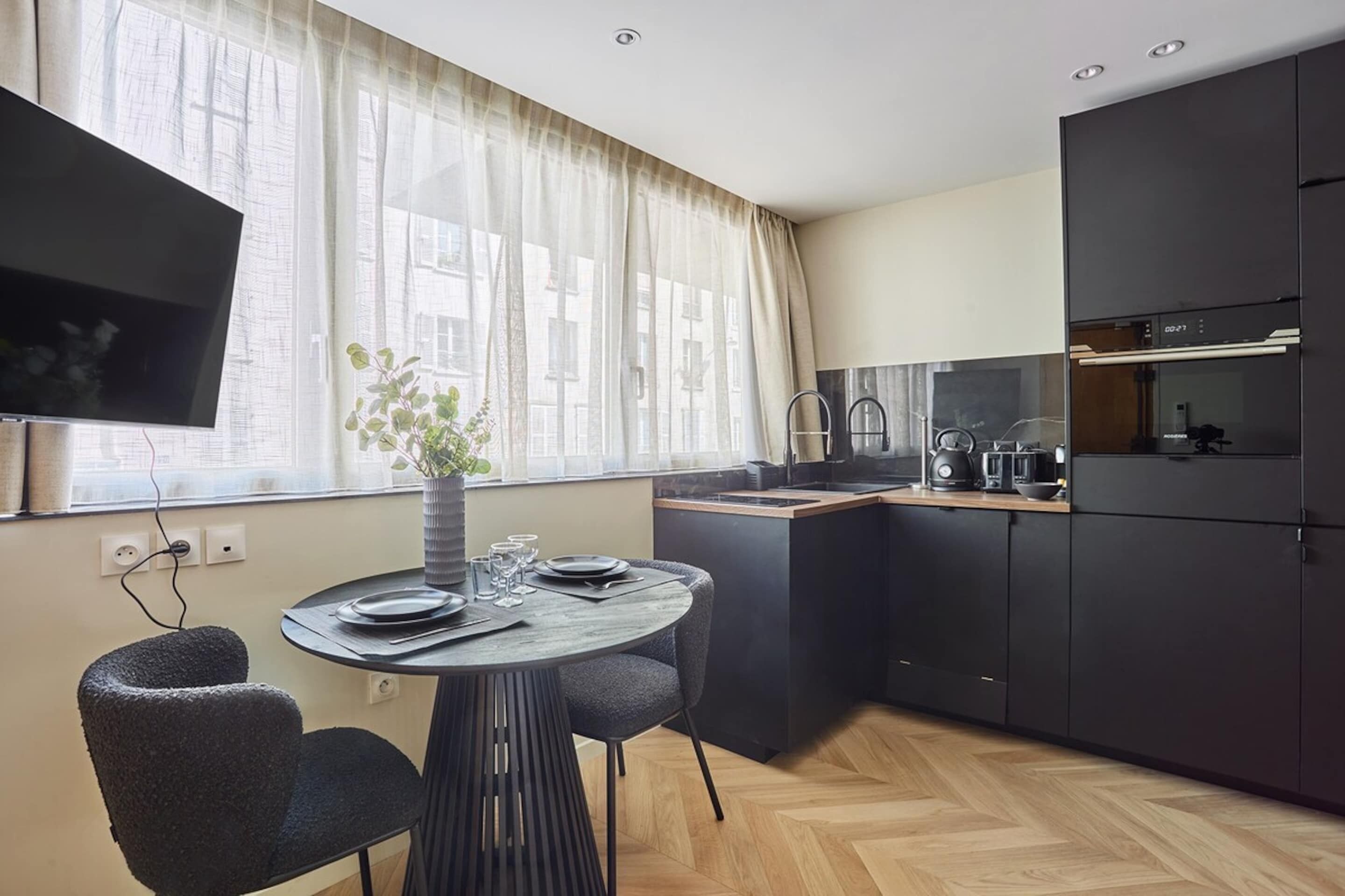 A modern kitchen with a dining area, a flat-screen TV, and a potted plant.