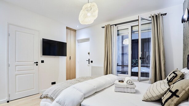 A bedroom with a bed, a TV mounted on the wall, a window with curtains, and a door. - Superb New Studio in the Center of Paris/opera (Paris)