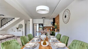 A dining room with a wooden table set for six, surrounded by green upholstered chairs. A large wall clock and a decorative vase with flowers are visible.